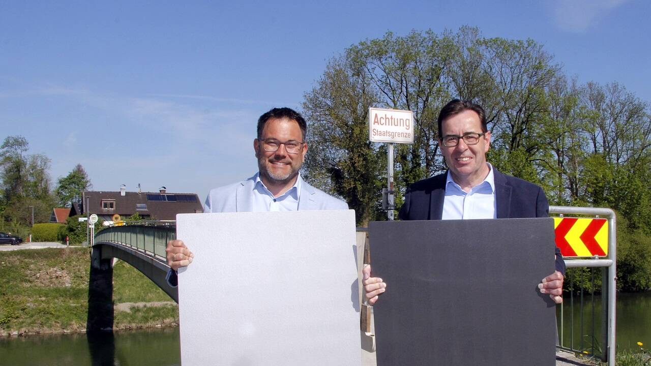 Die Bürgermeister der Gemeinden Ainring (Bayern) und Wals-Siezenheim (Flachgau), Martin Öttl und Andreas Hasenöhrl, vor dem Grenzsteg mit Farbmustern, mit denen der neue Steg gestaltet wird.  