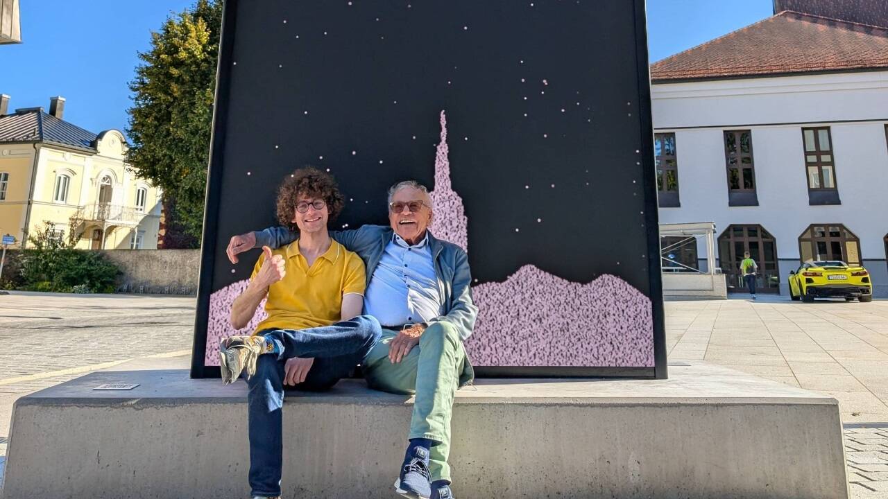 Christof Babinsky sitzt – hier mit seinem Vater Horst – auf dem Sockel seines Digitalen Monolithen. Auf der Glas- und LED-Fläche dahinter ist Babinskys Werk „Pixelrain“ zu sehen.  