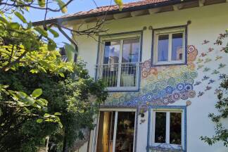 Der Regenbogen reicht von der Terrasse ins erste Stockwerk des Wohnhauses hinauf. Aus seinem oberen Ende fliegen Vögel heraus.  